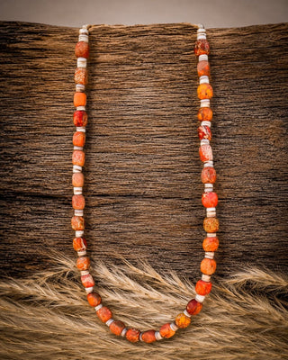 A beaded necklace made of orange and red marbled beads separated by white spacer beads, displayed on a rustic brown wooden surface with tan fur at the bottom.