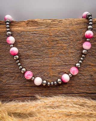 A beaded necklace with alternating pink and white marbled round beads, white beads, and dark metallic gray beads, displayed on a rustic wooden block with dried grass in the foreground.