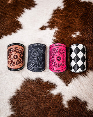 Four leather scarf slides arranged in a row on a brown and white cowhide background. The slides feature different designs: a tan floral tooled design, a dark gray embossed floral design, a bright pink floral tooled design, and a black and white harlequin diamond pattern.
