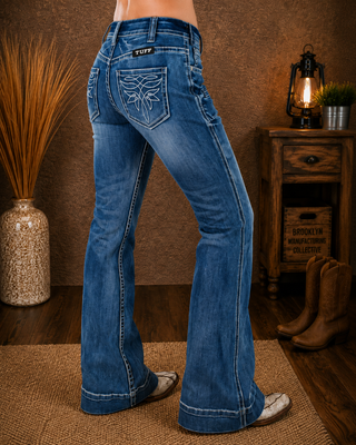 A person wearing blue western flare jeans with embroidered detailing on the back pocket, shown from the waist down in a rustic indoor setting with a wooden cabinet, lantern, and cowboy boots.