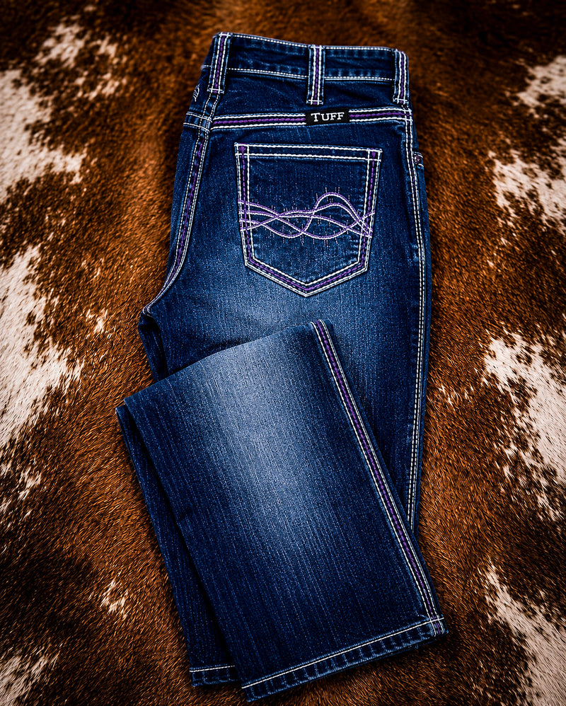 A pair of blue denim jeans laid flat on a brown and white cowhide rug. The jeans feature purple contrast stitching and a decorative purple embroidered pocket detail on the back pocket.