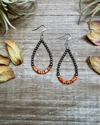 Teardrop-shaped earrings with orange beads on a wooden surface with dried flowers.