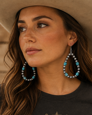 A woman wearing a beige cowboy hat and a dark graphic t-shirt with a longhorn skull design, featuring large turquoise and silver beaded teardrop earrings. She is posed against a rustic wooden wall.