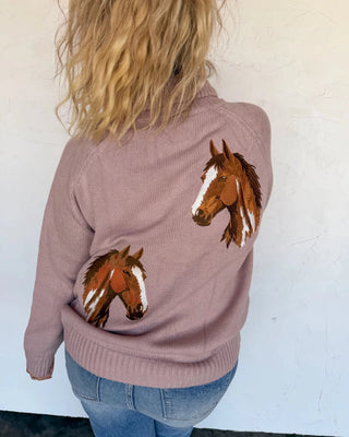 A woman viewed from behind wearing a dusty mauve knit sweater with embroidered brown horse heads on the back, paired with blue jeans against a white wall.
