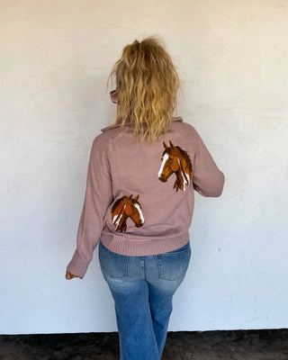 A woman viewed from behind wearing a dusty rose pink knit sweater with embroidered brown and white horse heads on the back, paired with blue jeans, standing against a white wall.