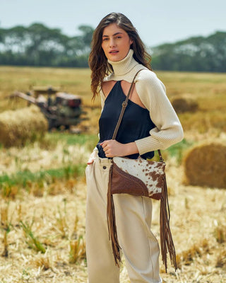 Woman standing in a field with hay bales, holding a cowhide bag.
