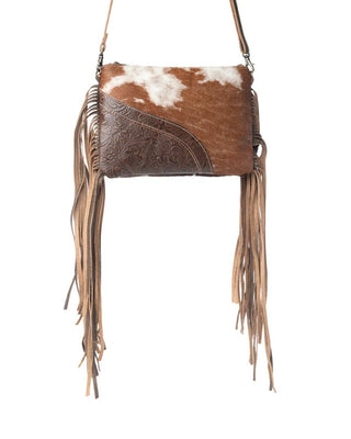 A western-style crossbody bag featuring a brown and white cowhide flap, an embossed brown leather panel, and long tan leather fringe, shown against a plain white background.
