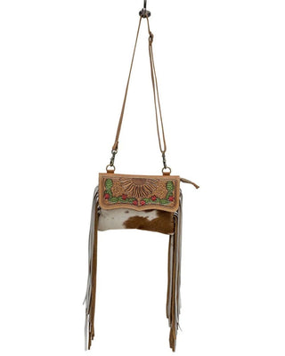 A western-style fringe crossbody bag with a tan leather flap featuring a sunburst tooling design and colorful beadwork, a brown and white cowhide body, and long tan leather fringe, shown against a plain white background.