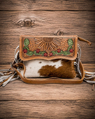A hand-tooled leather clutch bag with a brown and white cowhide body, featuring a sunflower and cactus design on the flap, set against a rustic wooden background.