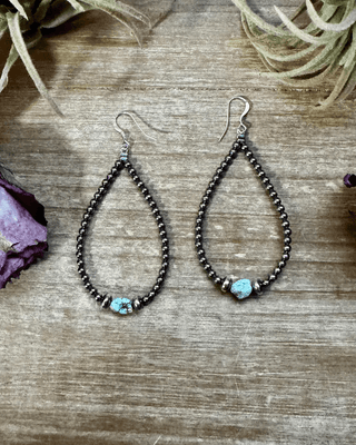 Teardrop-shaped beaded earrings with turquoise stones on a wooden surface with dried flowers.