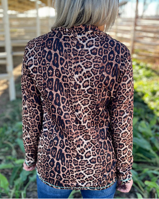 A woman viewed from behind wearing a leopard print long-sleeve top and blue jeans in an outdoor setting with a wooden structure and greenery in the background.