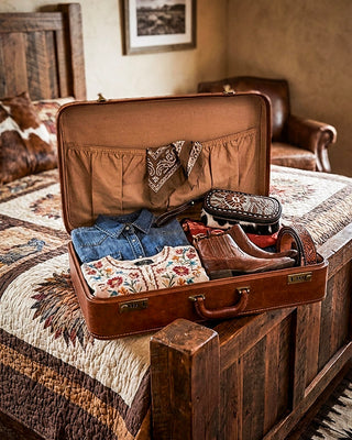 An open brown leather suitcase on a rustic wooden bed, packed with folded clothing, a bandana, a small patterned pouch, and a pair of brown leather boots in a warm, western-style bedroom setting.