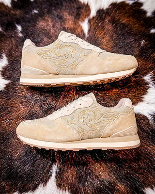 A pair of beige suede sneakers with tonal embroidered swirl patterns, displayed on a brown and white cowhide rug.