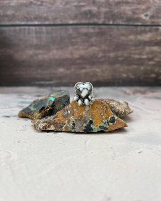 A silver-toned heart-shaped ring with white stones, displayed on rough natural rocks against a rustic wood and light surface background.