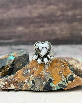 A silver ring with a heart-shaped white stone and smaller white stones around it, displayed on a textured orange and brown rock with other rocks in the background.