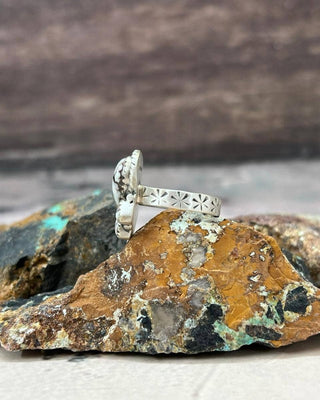 A sterling silver ring with a textured, organic-shaped setting and a band featuring starburst cutouts, displayed on a piece of raw turquoise and copper mineral rock.