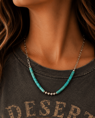 A close-up of a woman wearing a turquoise and silver beaded necklace with a silver chain, paired with a dark gray graphic t-shirt featuring a desert sunset design.