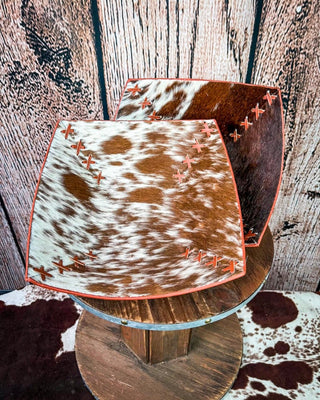 Two cowhide-patterned square trays with reddish-brown stitched edges, displayed on a rustic wooden spool against a wood plank backdrop.