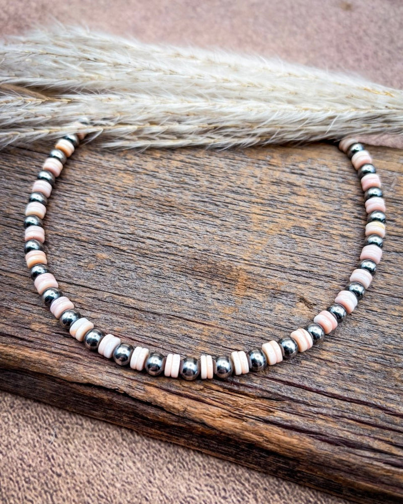 A beaded necklace with alternating light pink and black marbled beads, displayed on a rustic wooden surface with dried grass in the background.