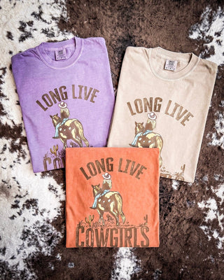 Three folded graphic T-shirts in purple, beige, and orange are arranged on a brown and white cowhide rug. Each shirt features a cowboy riding a cowgirl with the words 'LONG LIVE COWGIRLS' printed above and below the illustration.