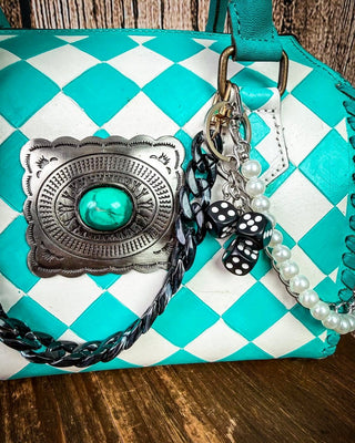 A turquoise and white checkered leather handbag with a large silver concho featuring a turquoise center, adorned with braided black and white strap, pearl beads, and a black-and-white polka dot bow charm on a wooden surface.