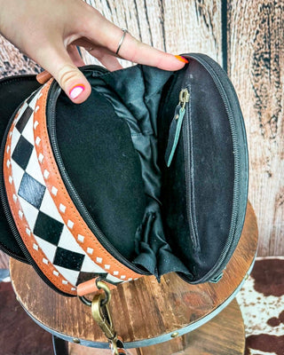 A close-up of a hand holding open a small black zippered clutch or cosmetic bag, showing the black interior lining and a black-and-white harlequin patterned exterior trim with tan leather edging. The bag is photographed over a round wooden surface against a rustic wood plank background.