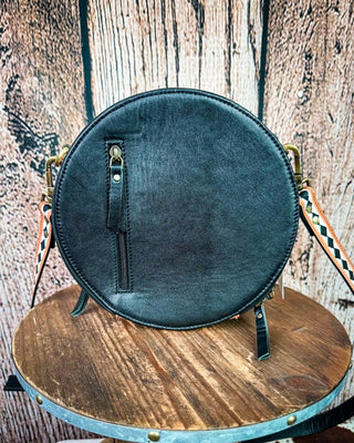 A round black leather crossbody bag with brass hardware and a patterned strap, displayed on a rustic wooden table against a wood plank background.