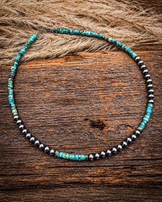 A beaded necklace with turquoise-blue and black beads, arranged in a circle on a rustic wooden surface with dried grass in the background.