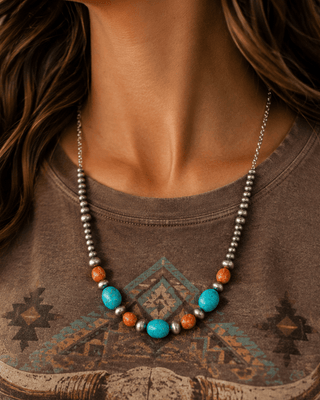 A close-up of a woman wearing a bohemian-style beaded necklace with turquoise, orange, and silver beads over a brown graphic t-shirt featuring a bull skull design.
