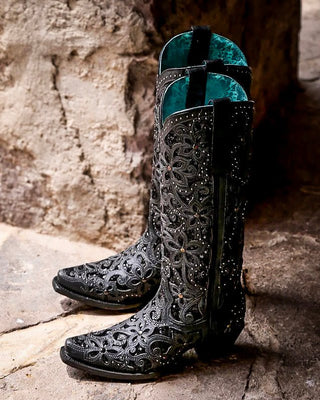 A pair of black western cowboy boots with intricate floral cutout embroidery and silver rhinestone embellishments, standing on a rustic stone surface against a textured stone wall.