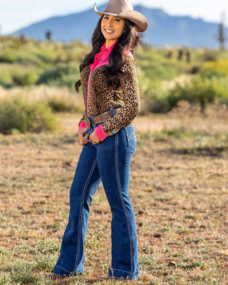 A woman stands outdoors in a dry grassy field wearing a tan cowboy hat, a leopard-print jacket over a bright pink top, blue jeans, and a brown belt, with mountains blurred in the background.