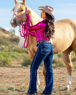 A woman in a cowboy hat, shiny pink long-sleeve top, and blue jeans stands beside a light tan horse outdoors, holding the horse's reins.