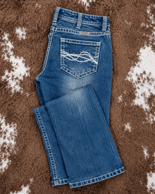 A pair of medium-wash blue jeans laid flat on a brown and white cowhide rug. The back pocket features a distinctive white embroidered wave pattern.