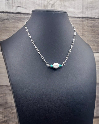 A silver paperclip chain necklace with a central white pearl bead flanked by two small turquoise blue beads, displayed on a black jewelry bust against a rustic wood background.
