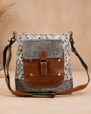 A rustic crossbody bag with a gray patterned fabric body, a brown faux-leather front pocket with a buckle closure, and a matching brown strap, set against a warm beige background.