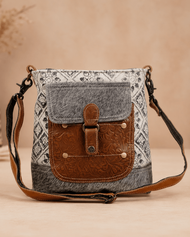A rustic crossbody bag with a gray patterned fabric body, a brown faux-leather front pocket with a buckle closure, and a matching brown strap, set against a warm beige background.