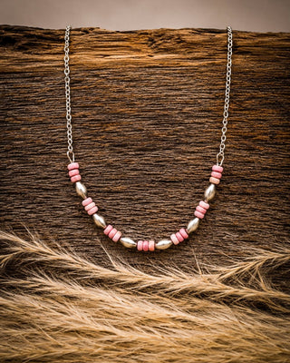 A silver chain necklace with alternating pink and metallic silver beads, displayed on a rustic dark wood background with dried grass in the foreground.