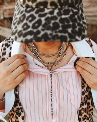 A close-up lifestyle shot of a person wearing a leopard print baseball cap with 'Bless To Me' text, a pink and white striped button-down shirt, and a matching leopard print vest. The person is wearing multiple layered silver necklaces and holding the collar open.