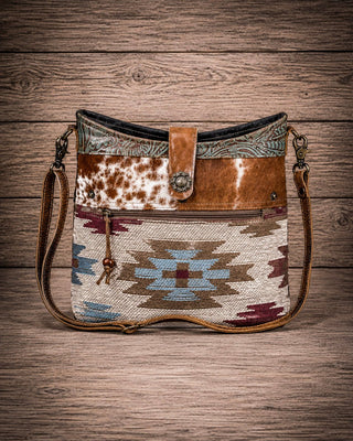 A western-style crossbody bag with a combination of cowhide, patterned leather, and a Southwestern geometric woven textile panel, displayed on a rustic wooden plank background.