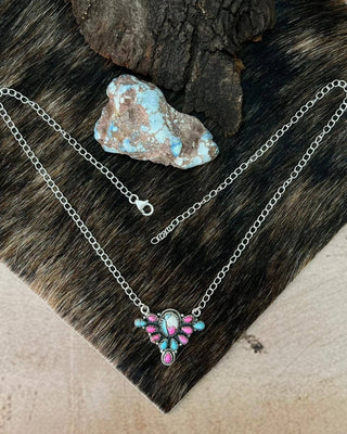 A silver chain necklace with a decorative pendant featuring turquoise and pink stones, displayed on dark animal fur with a small blue stone prop.