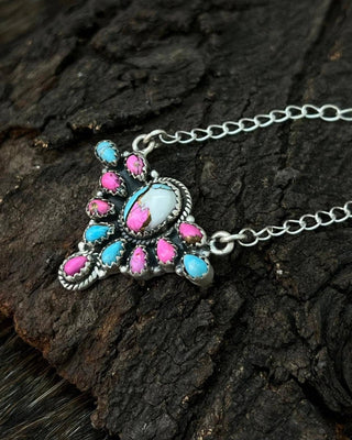 A silver-toned pendant necklace with a Southwestern-style design, featuring a large white oval stone, a central pink stone, and smaller pink and turquoise blue stones, displayed on a textured dark wood background.