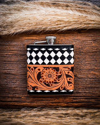 A small rectangular flask with a silver metal cap, decorated with a black-and-white diamond pattern on the upper half and a carved orange floral scroll design on the lower half, displayed on a rustic wooden surface with tan fur around it.