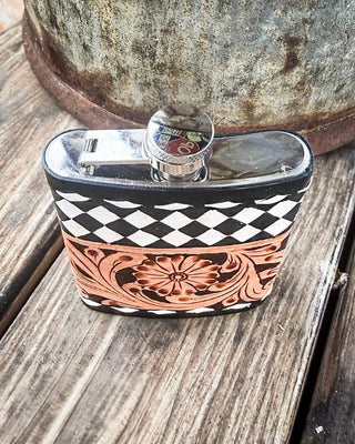A small rectangular metal flask with a black and white checkerboard pattern and a copper-toned floral tooling design on the front, resting on a weathered wooden surface with a rusty metal container in the background.