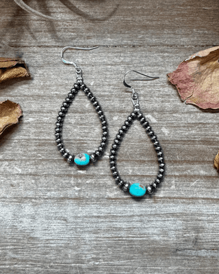 Teardrop-shaped beaded earrings with turquoise beads on a wooden surface with dried leaves.