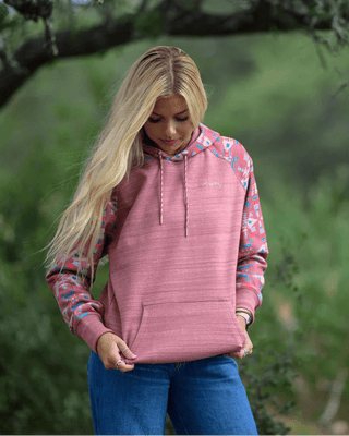 A woman wearing a dusty pink pullover hoodie with floral patterned sleeves, standing outdoors in a grassy, wooded setting.