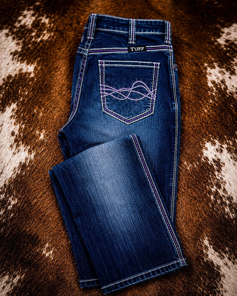 A pair of blue denim jeans laid flat on a brown and white cowhide rug. The jeans feature purple contrast stitching and a decorative purple embroidered pocket detail on the back pocket.