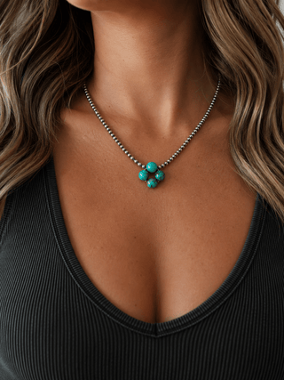 A close-up of a woman wearing a silver beaded chain necklace with a central cluster of turquoise-colored beads, paired with a black ribbed V-neck top.