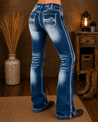 Rear view of a person wearing blue western flare jeans with white contrast stitching and decorative stitching on the back pockets, standing in a rustic room with cowboy boots, a wooden cabinet, and a lamp in the background.