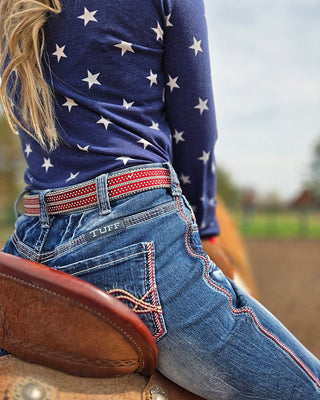 A close-up lifestyle shot of a person sitting on a brown leather saddle outdoors, wearing a navy blue long-sleeve shirt with white stars, blue jeans with red contrast stitching and embroidery, and a red patterned belt. The brand name 'TUFF' is visible on the back pocket.