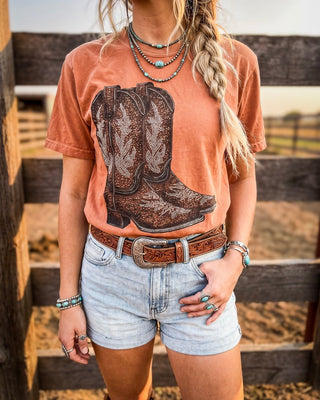 A woman wearing a faded rust t-shirt with a large brown cowboy boot graphic, styled with light-wash denim shorts, a brown leather belt, and layered turquoise jewelry, standing in front of a wooden fence.